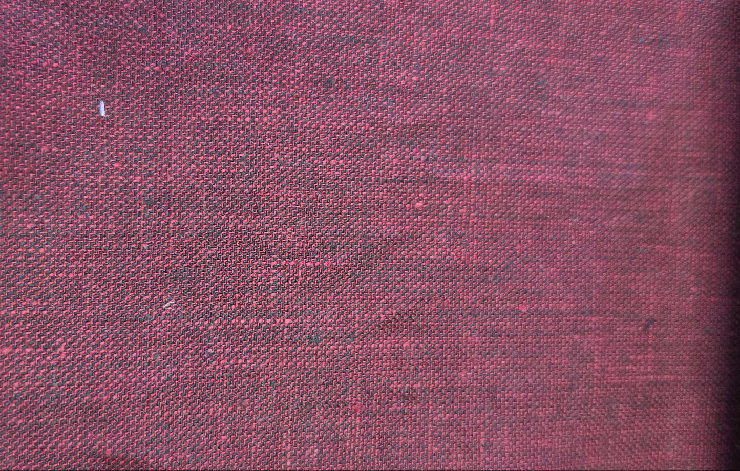 A close-up of a textured fabric swatch in a deep burgundy red, showing a woven linen-like pattern, with a beige zigzag edge and a light wood block visible on the left.