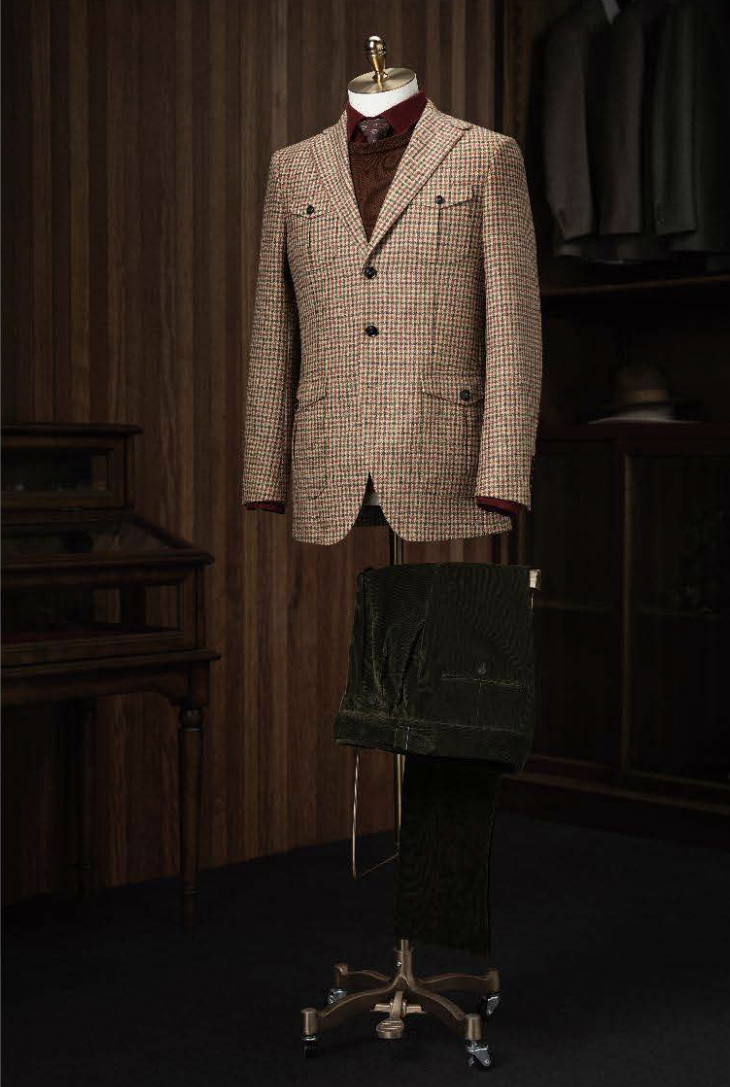 A suit jacket and trousers displayed on a mannequin in a room with wood paneling, featuring a burgundy/green check tweed pattern.