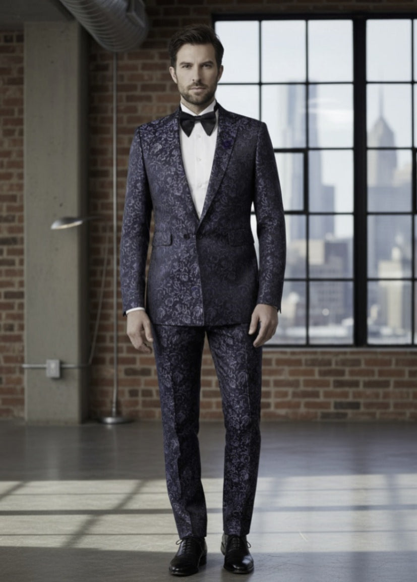 Man wearing a dark patterned suit with a white shirt and black bow tie in an urban setting.