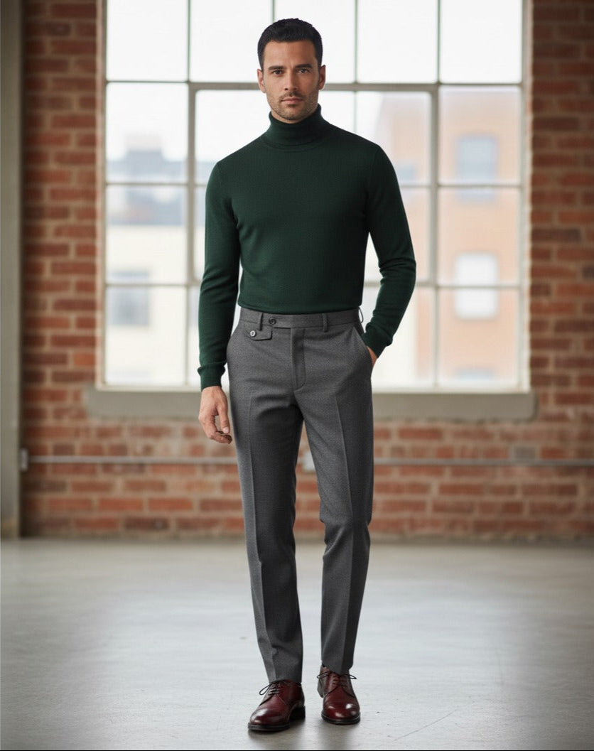 Man wearing a green turtleneck and gray pants standing in front of a brick wall with large windows.