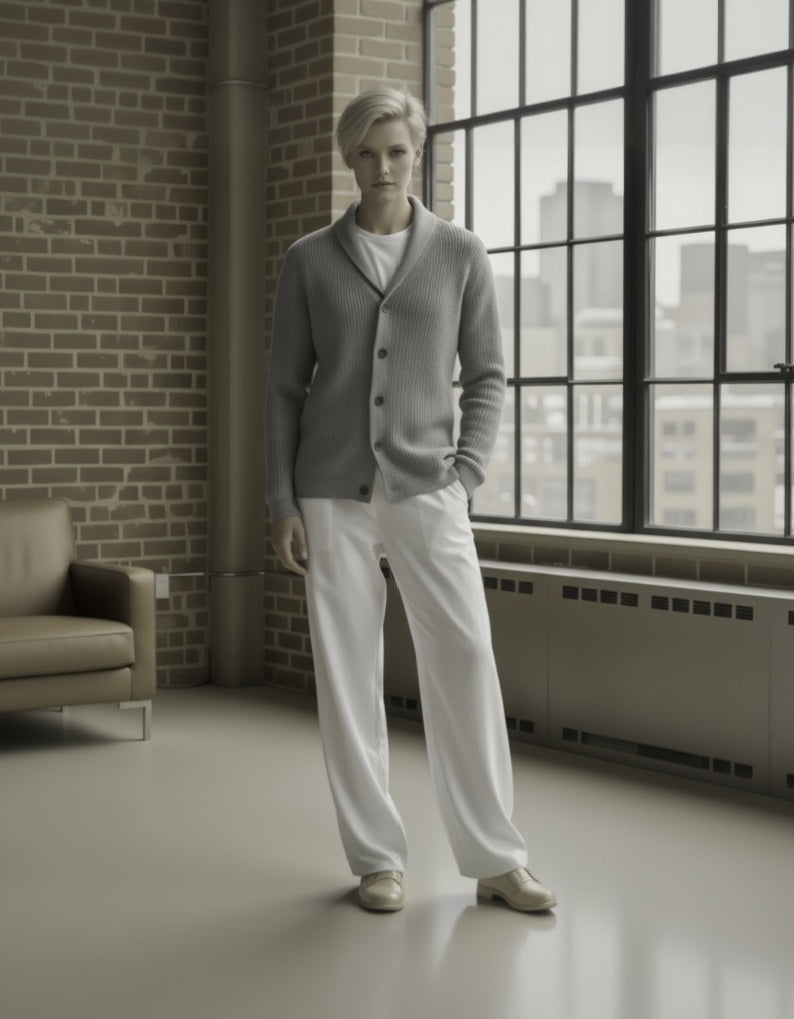 Person wearing a gray cardigan and white pants standing in a room with large windows and brick wall.