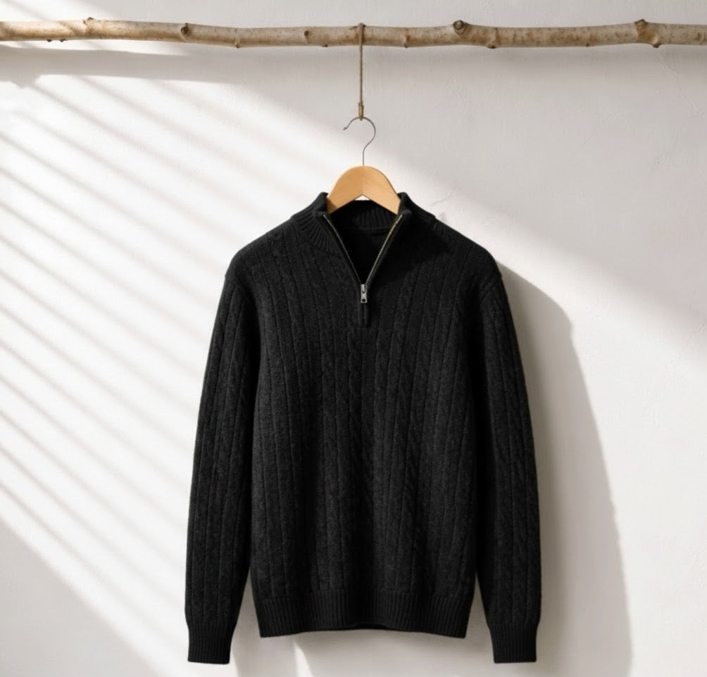 A black cable-knit quarter-zip sweater hanging on a wooden hanger from a rustic branch, set against a white wall with diagonal shadow patterns.