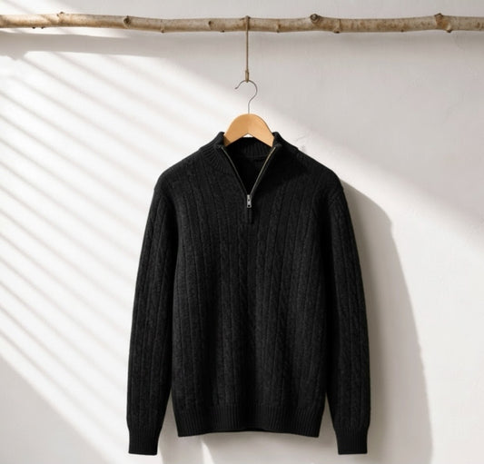 A black cable-knit quarter-zip sweater hanging on a wooden hanger from a rustic branch, set against a white wall with diagonal shadow patterns.