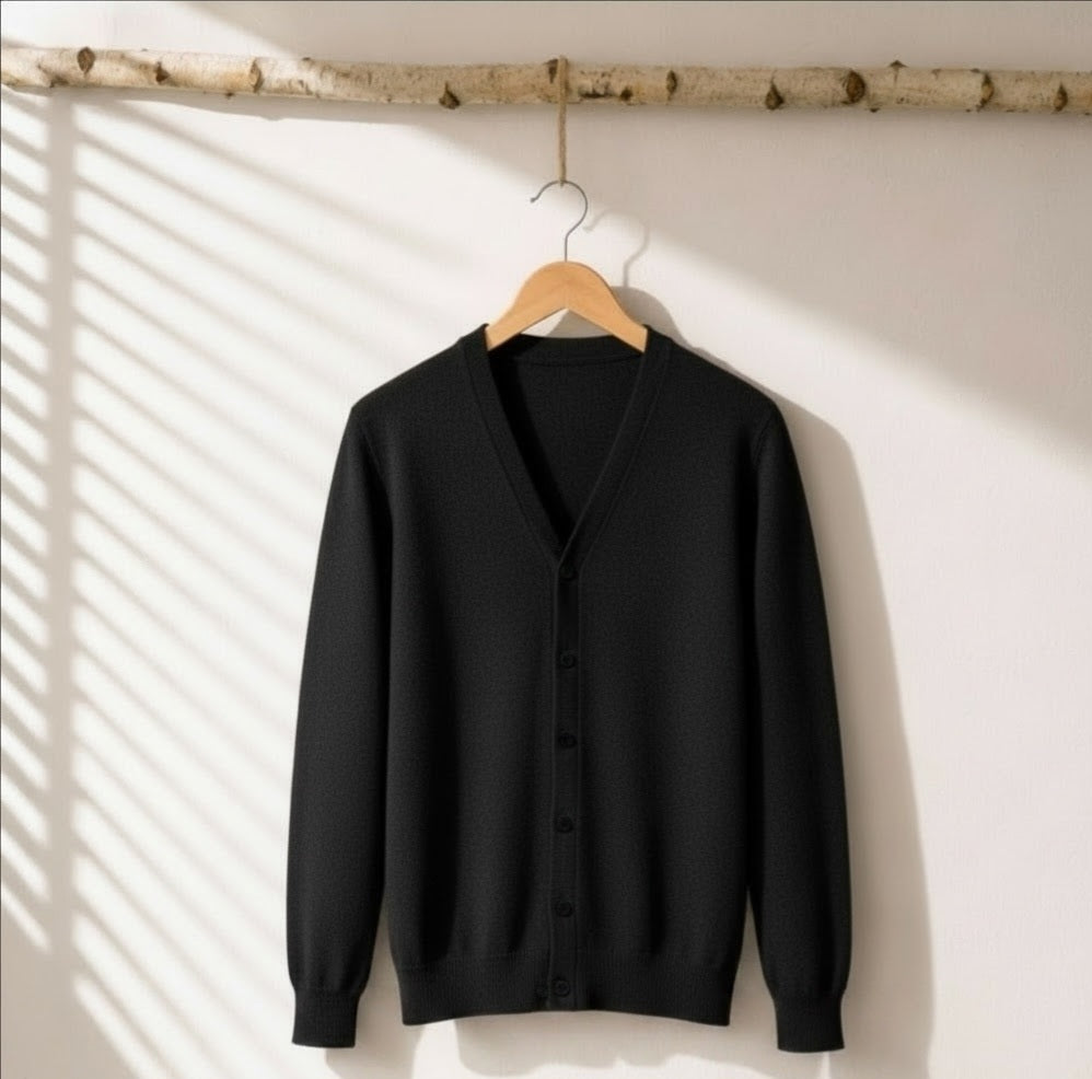 A black knit cardigan sweater with a V-neck and button-front closure hangs on a wooden hanger from a birch branch, set against a white wall with soft window shadow patterns.