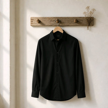 A black long-sleeve button-up shirt hanging on a wooden hanger from a rustic wooden wall hook, set against a textured off-white wall with soft window light shadows.