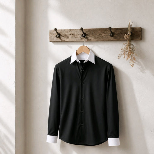 A black long-sleeve button-up shirt with a white pointed collar and white cuff placket, hanging on a wooden hanger from a rustic wall-mounted wooden coat rack with dried flowers.