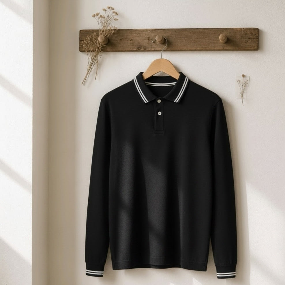 A black long-sleeve polo shirt with white striped collar and cuffs, hanging on a wooden hanger from a rustic wooden peg rail against a warm off-white wall.