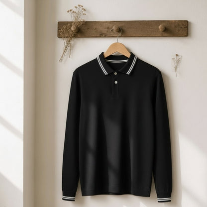 A black long-sleeve polo shirt with white striped collar and cuffs, hanging on a wooden hanger from a rustic wooden peg rail against a warm off-white wall.