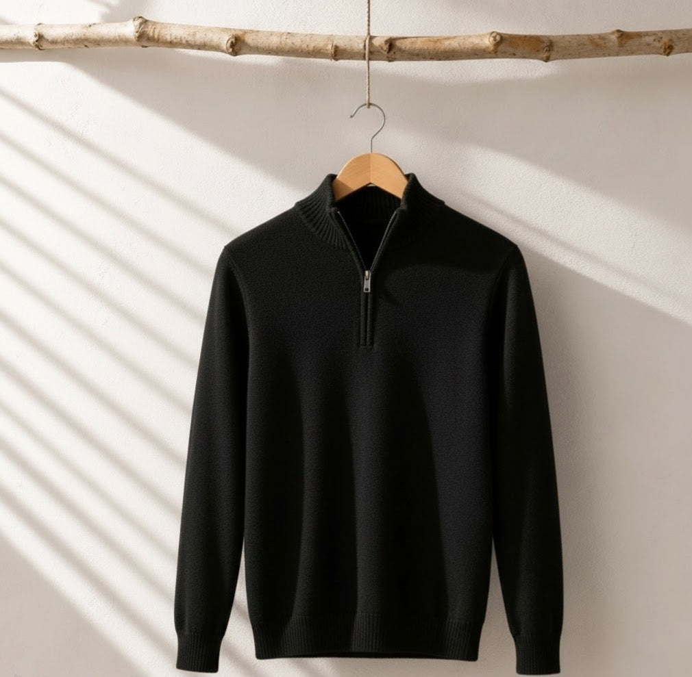 A black long-sleeve quarter-zip knit sweater hanging on a wooden hanger from a natural branch, set against a white wall with soft diagonal shadow patterns.