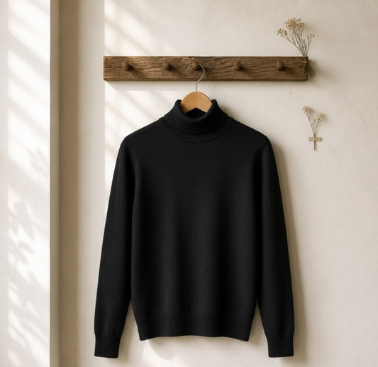 A black ribbed turtleneck sweater hanging on a wooden hanger from a rustic wooden wall rack, with dried flowers on the wall and soft window shadows casting across the scene.