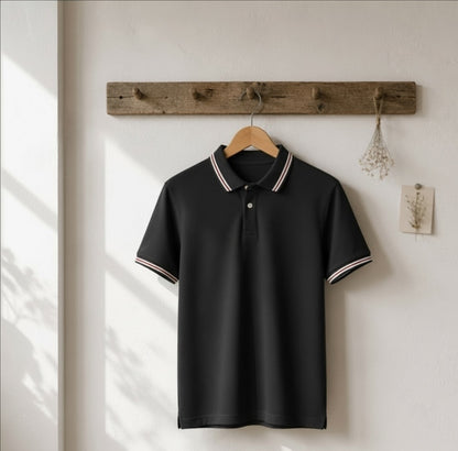 A black short-sleeve polo shirt with white and navy striped trim on the collar and sleeve cuffs, hanging on a wooden hanger from a rustic wooden wall rack against a white wall.