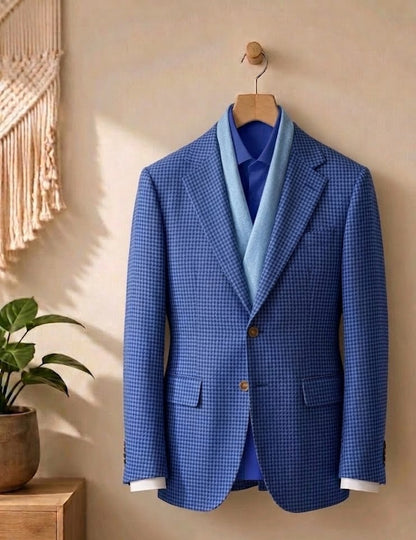 A blue and white checkered blazer jacket hanging on a wooden hanger against a beige wall, styled with a light blue knit sweater vest, a royal blue dress shirt, and white dress cuffs, with a potted plant and macrame wall hanging in the background.