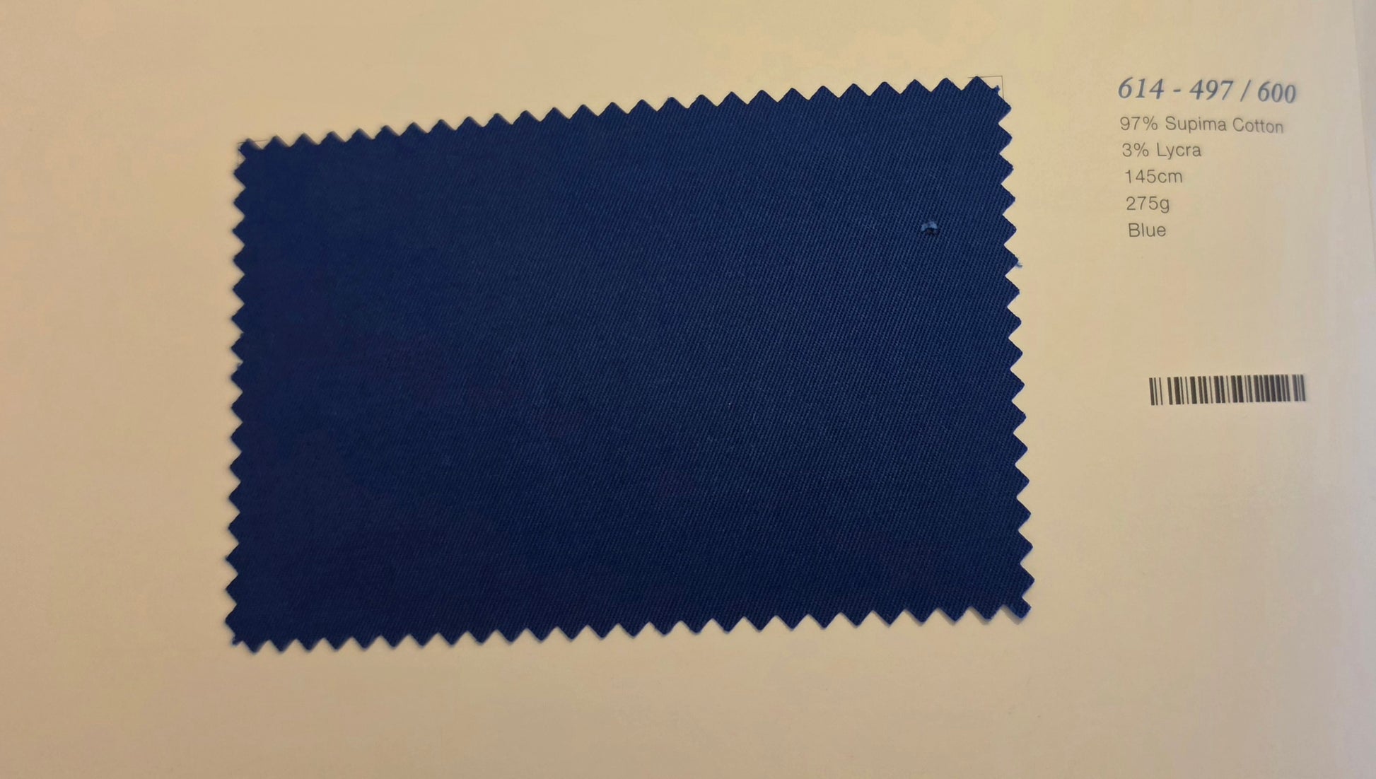 A blue fabric swatch card with serrated edges on a light beige background, printed with color and material details including '614 - 497 / 600' and 'Blue'.
