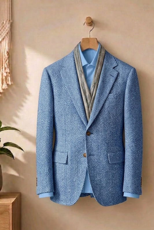 A blue herringbone blazer jacket hanging on a wooden hanger against a warm beige wall, styled with a light blue dress shirt and a gray neck scarf. A wooden side table with a green plant is visible on the left.