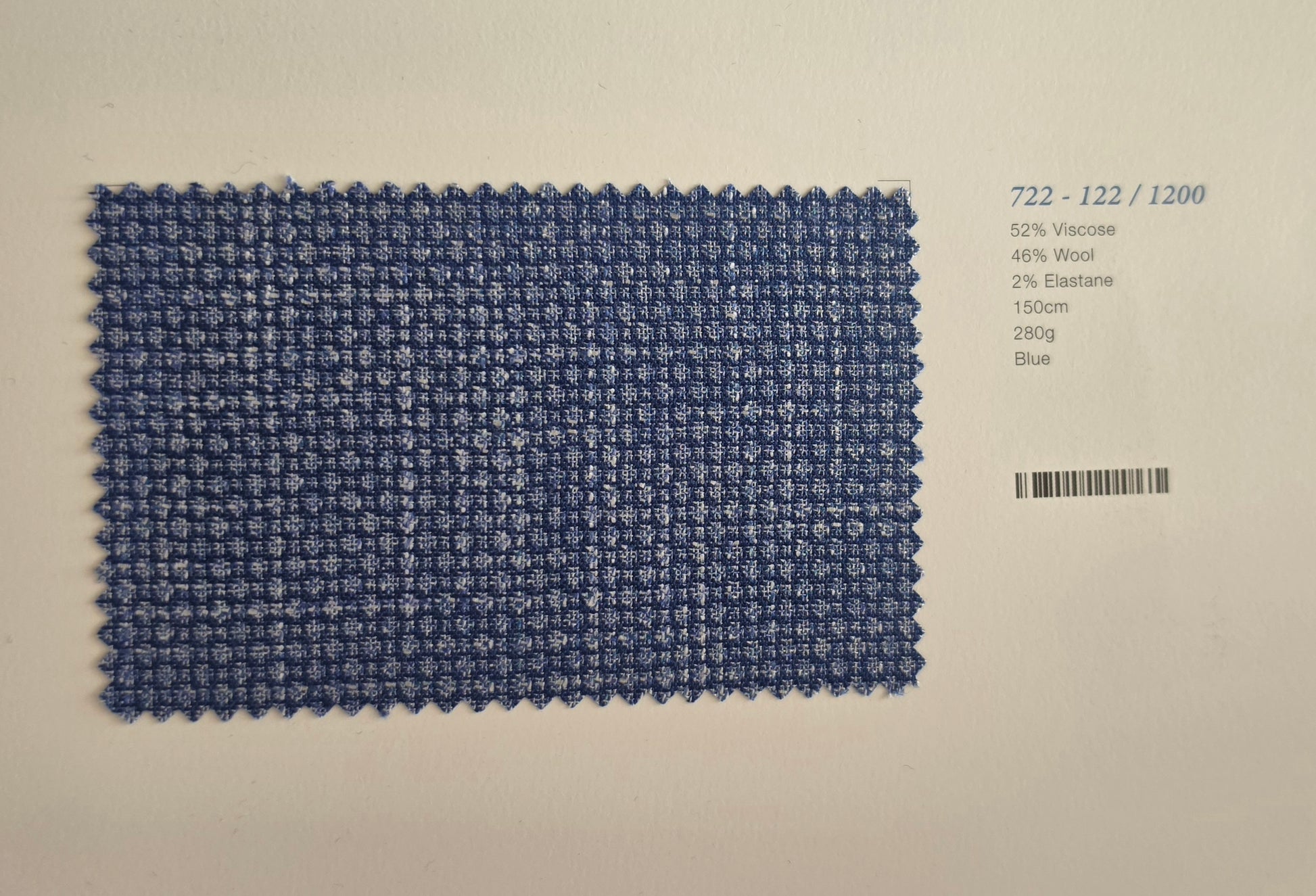 A blue woven fabric swatch card with a barcode and material details printed on the right side, set against a plain off-white background.