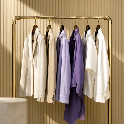A brass clothing rack holding six shirts or lightweight blouses in different colors: off-white, cream, light purple, dark purple, white, and light gray. The rack is set against a beige vertically slatted wall with warm sunlight and shadows.