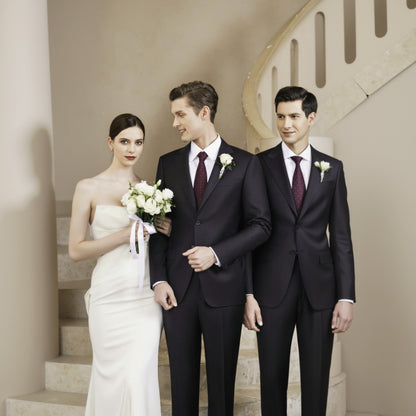 A bride in a white dress holding a bouquet, standing next to a groom in a dark grey pinstripe suit with a notched lapel and a working cuff, with another man in a black suit looking on.