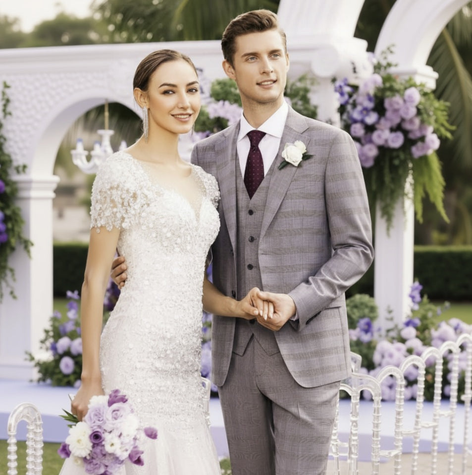 A bride in a white floral gown and a groom in a grey three-piece Italian suit with merino wool details, standing together at a wedding.