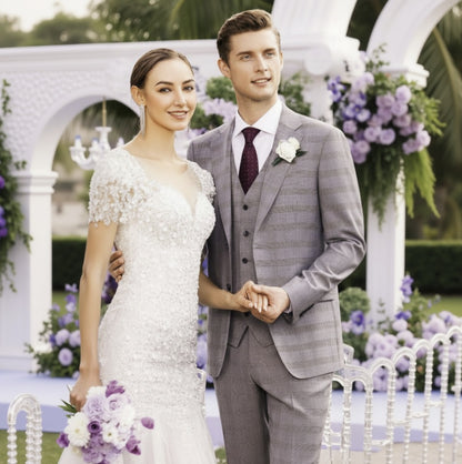 A bride in a white floral gown and a groom in a grey three-piece Italian suit with merino wool details, standing together at a wedding.