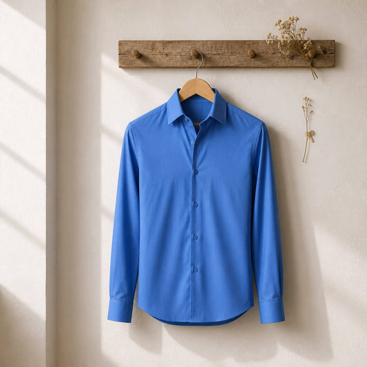 A bright blue long-sleeve button-up dress shirt hanging on a wooden hanger from a rustic wooden wall rack, set against a warm beige textured wall with soft window light shadows.