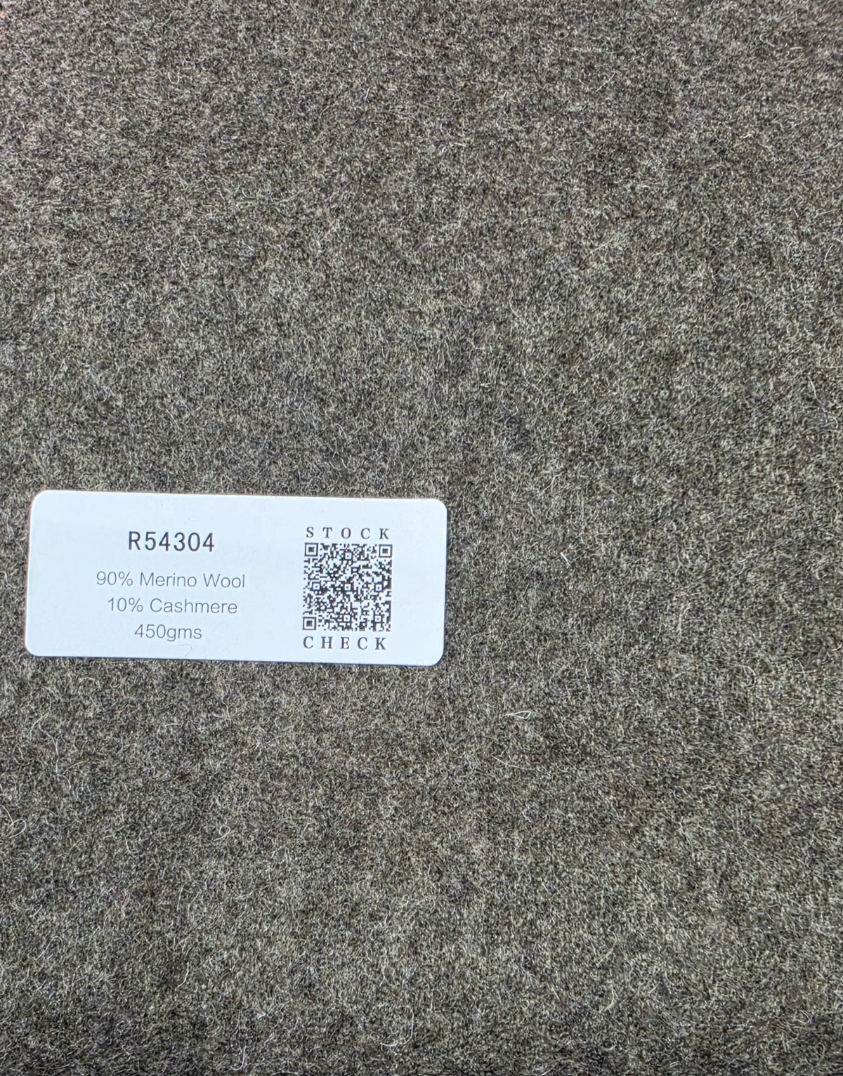 A close-up of a gray textured fabric swatch with a white product label that reads '90% Merino Wool, 10% Cashmere, 450gms' and includes a QR code.
