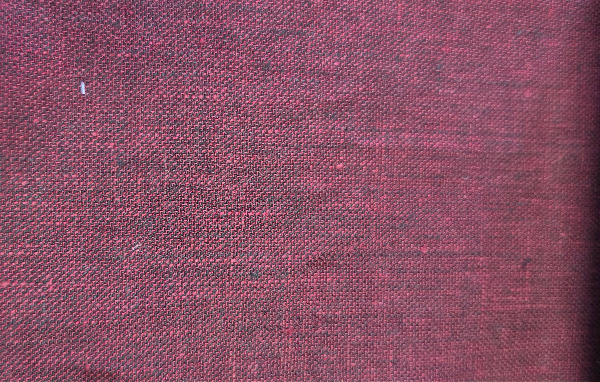 A close-up of a textured fabric swatch in a deep burgundy red, showing a woven linen-like pattern, with a beige zigzag edge and a light wood block visible on the left.