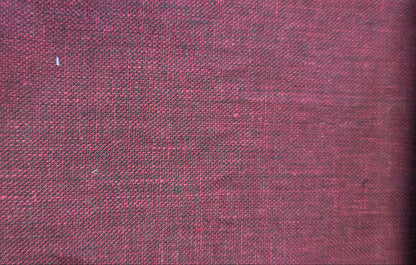 A close-up of a textured fabric swatch in a deep burgundy red, showing a woven linen-like pattern, with a beige zigzag edge and a light wood block visible on the left.