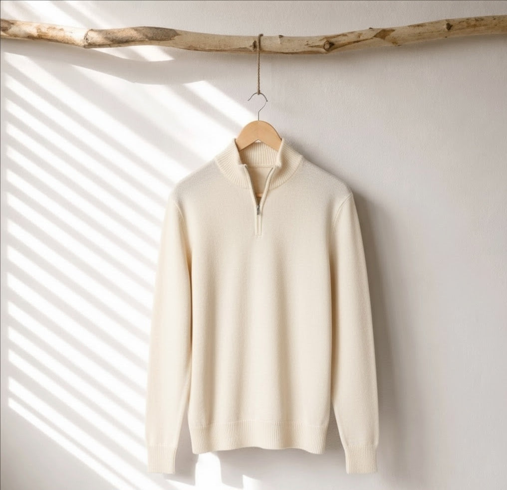 A cream-colored half-zip knit sweater hanging on a wooden hanger from a rustic branch, set against a white wall with soft diagonal window shadow patterns.