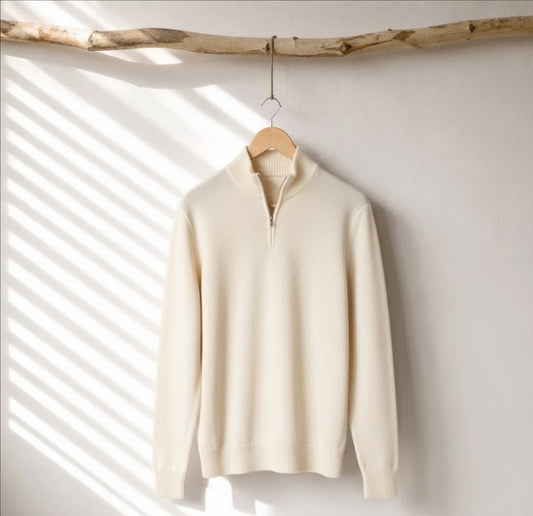 A cream-colored half-zip knit sweater hanging on a wooden hanger from a rustic branch, set against a white wall with soft diagonal window shadow patterns.