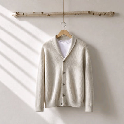A cream-colored ribbed knit cardigan sweater with a shawl collar, hanging on a wooden hanger from a birch branch against a warm off-white wall with soft diagonal shadows.