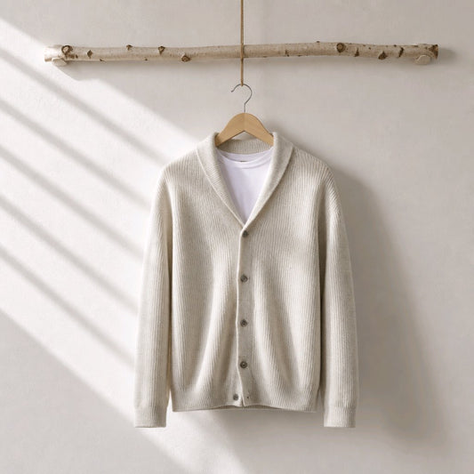 A cream-colored ribbed knit cardigan sweater with a shawl collar, hanging on a wooden hanger from a birch branch against a warm off-white wall with soft diagonal shadows.