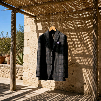 A dark charcoal gray textured blazer with a subtle plaid pattern hangs on a wooden hanger from a rustic thatched roof, set against a stone wall in warm, sunny outdoor light.