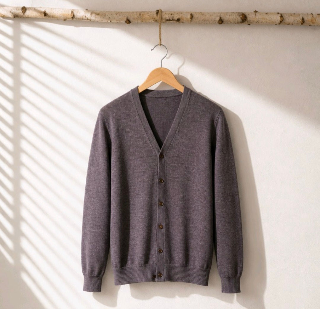A dark gray knit cardigan sweater with a V-neck and button-front closure, hanging on a wooden hanger from a birch branch against a white wall with soft window shadow patterns.
