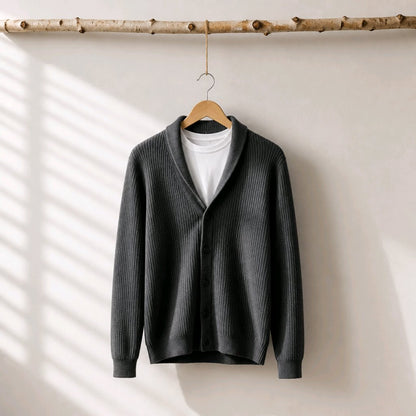 A dark gray ribbed cardigan sweater with a shawl collar, hanging on a wooden hanger from a natural birch branch. The cardigan is layered over a white crewneck t-shirt, set against a warm off-white wall with soft window shadow patterns.