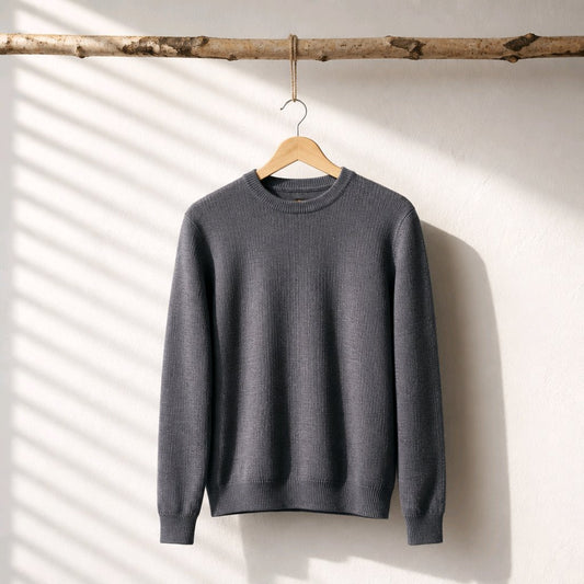 A dark gray ribbed knit crewneck sweater hanging on a wooden hanger from a natural wood branch, set against a white wall with soft diagonal window shadow patterns.
