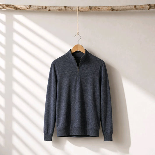 A dark heather gray quarter-zip pullover sweater hanging on a wooden hanger from a rustic birch branch, set against a white wall with soft diagonal window shadow patterns.