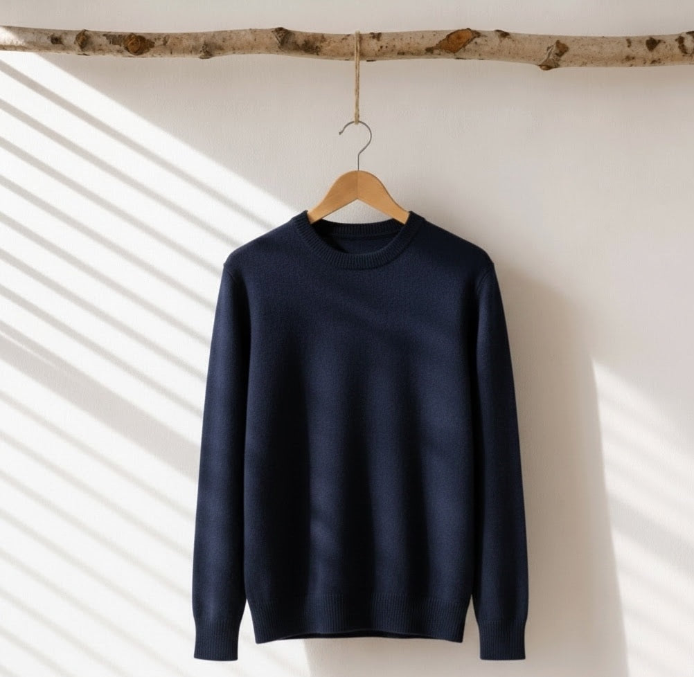 A dark navy blue crewneck sweater hanging on a wooden hanger from a birch branch, set against a white wall with soft diagonal window shadow patterns.