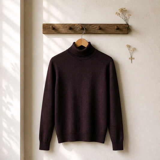 A dark plum purple knit turtleneck sweater hanging on a wooden hanger from a rustic wooden wall rack, with soft window shadow patterns on a textured off-white wall.