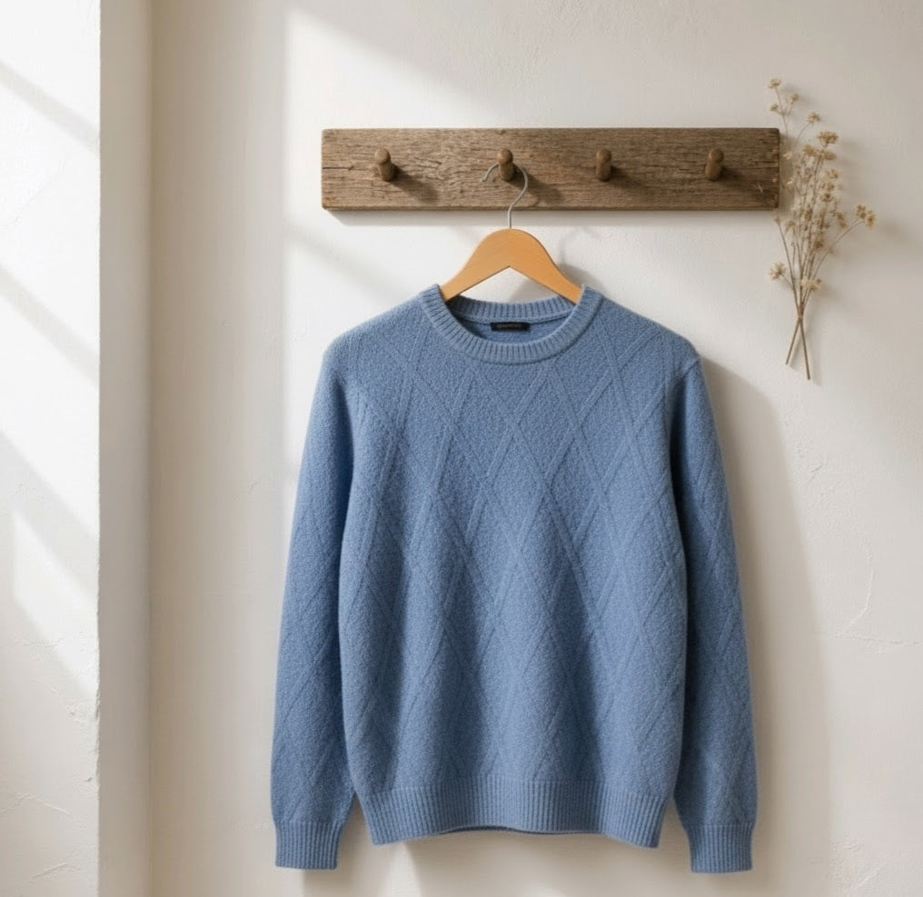 A dusty blue crewneck sweater with a diamond cable-knit pattern hanging on a wooden hanger from a rustic wooden wall hook.