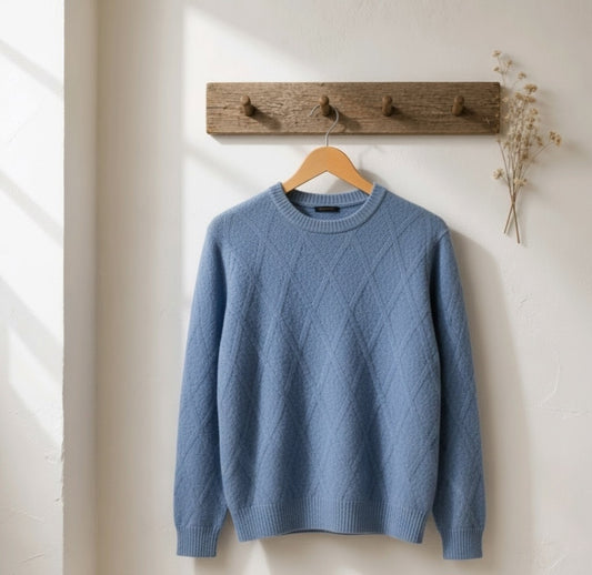 A dusty blue crewneck sweater with a diamond cable-knit pattern hanging on a wooden hanger from a rustic wooden wall hook.