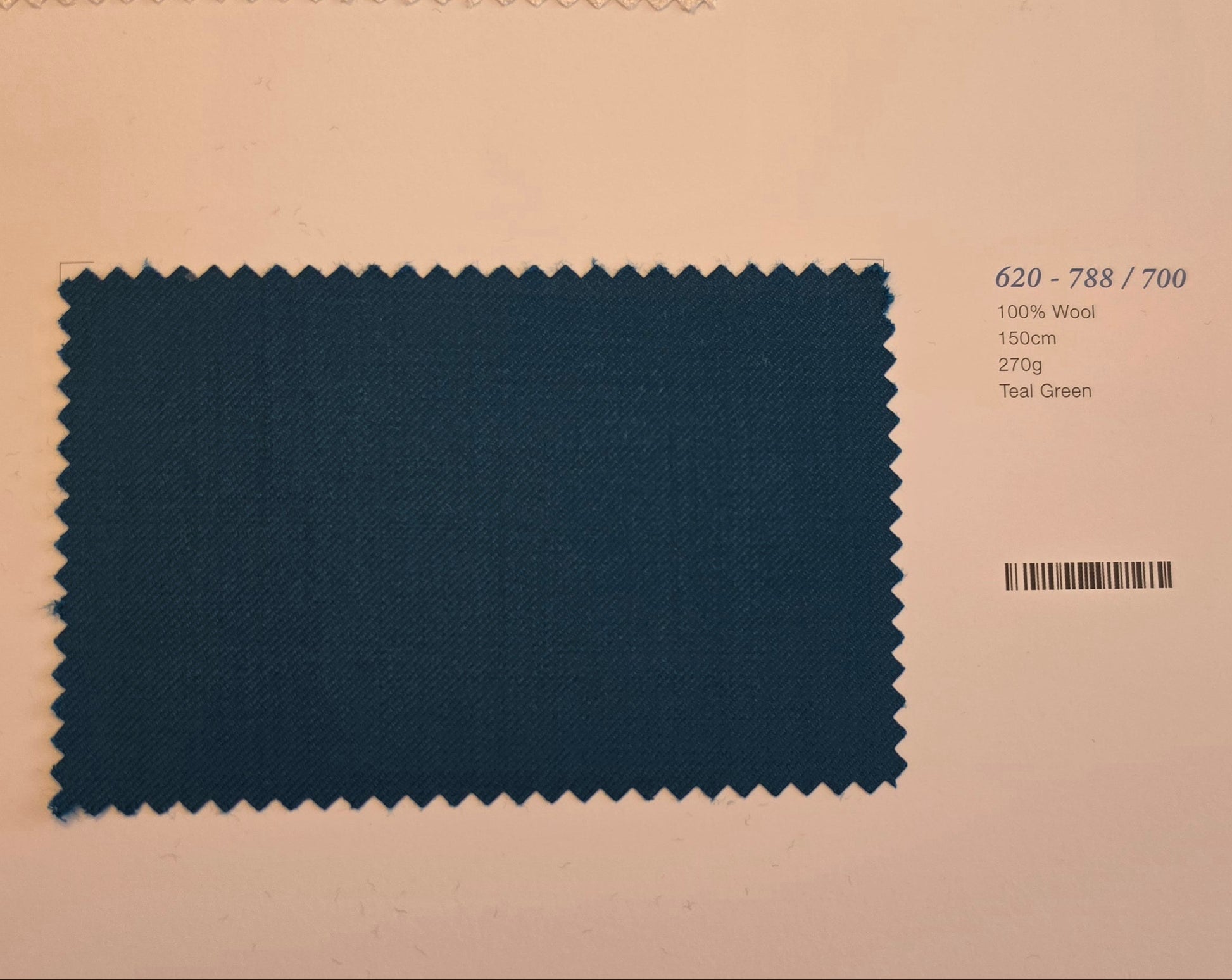 A fabric swatch card displaying a teal green wool sample with serrated edges, labeled with color code 620-788/700 and material details on a light beige background.