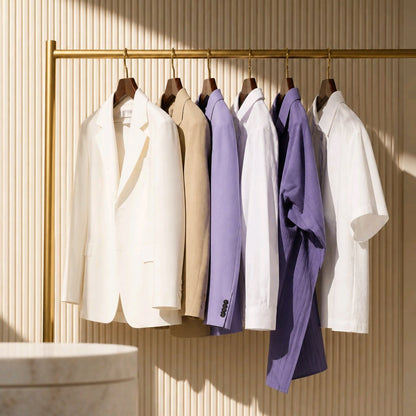 A gold clothing rack displaying a curated collection of men's apparel, including a white blazer, beige blazer, white shirt, lavender shirt, purple overshirt, and white short-sleeve shirt, set against a beige slatted wall.