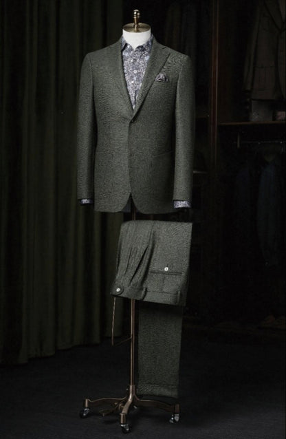 A green herringbone wool single-breasted jacket and matching wool trousers displayed on a mannequin, featuring a notched lapel, front buttons, and waistcoat.