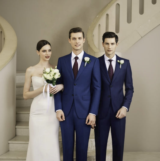 A group of three people, two men in blue suits and a woman in a white dress, standing on a staircase. The men are wearing ties, and the woman is holding a bouquet of flowers.