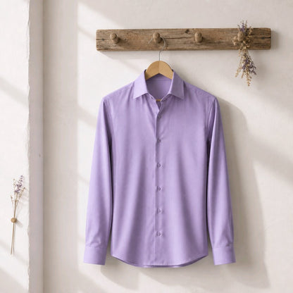 A lavender purple long-sleeve button-down dress shirt hanging on a wooden hanger from a rustic wooden wall hook, set against a warm off-white wall with soft sunlight shadows.