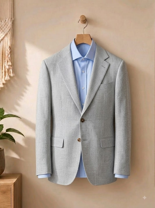A light Stone textured blazer jacket hanging on a wooden hanger against a warm beige wall, layered over a light blue dress shirt. A small potted plant and a macrame wall hanging are visible in the softly lit background.