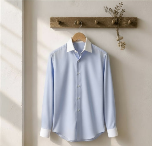 A light blue long-sleeve button-up dress shirt with a white collar and white cuffs, hanging on a wooden hanger from a rustic wooden wall hook.