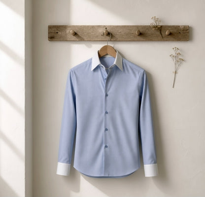 A light blue long-sleeve button-up dress shirt with a white collar and white cuffs, hanging on a wooden hanger from a rustic wooden wall rack against a textured off-white wall.