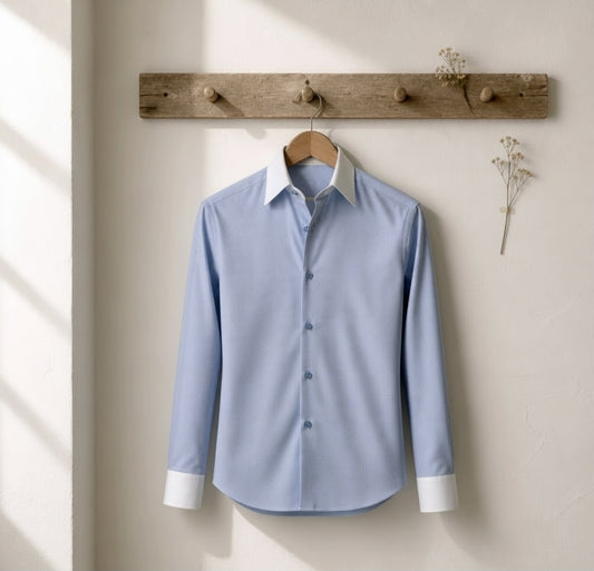 A light blue long-sleeve button-up dress shirt with a white collar and white cuffs, hanging on a wooden hanger from a rustic wooden wall rack against a textured off-white wall.