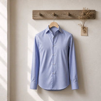 A light blue long-sleeve button-up shirt hanging on a wooden hanger from a rustic wooden wall hook, with a small vase of dried flowers to the right, set against a textured off-white wall with soft window shadows.